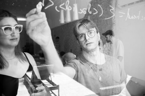 Another photo, this one looking through a window at Brooklyn Zelenka and Alex Good, who are collaborating on a design by writing notes on the window with a special pen. Again, the photo is dithered with big 1-bit black-and-white pixels.